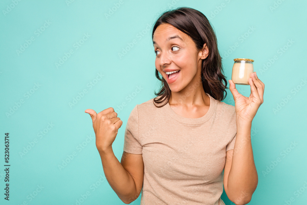 Young hispanic woman holding yogurt isolated on blue background points with thumb finger away, laughing and carefree. Young hispanic woman holding yogurt isolated on blue background points with thumb finger away, laughing and carefree.