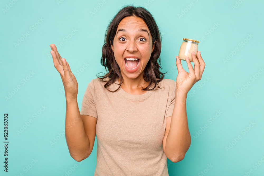 Young hispanic woman holding yogurt isolated on blue background receiving a pleasant surprise, excited and raising hands. Young hispanic woman holding yogurt isolated on blue background receiving a pleasant surprise, excited and raising hands.
