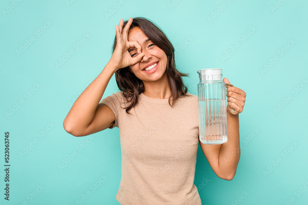 Young hispanic woman holding a water of jar isolated on blue background excited keeping ok gesture on eye. Young hispanic woman holding a water of jar isolated on blue background excited keeping ok gesture on eye.