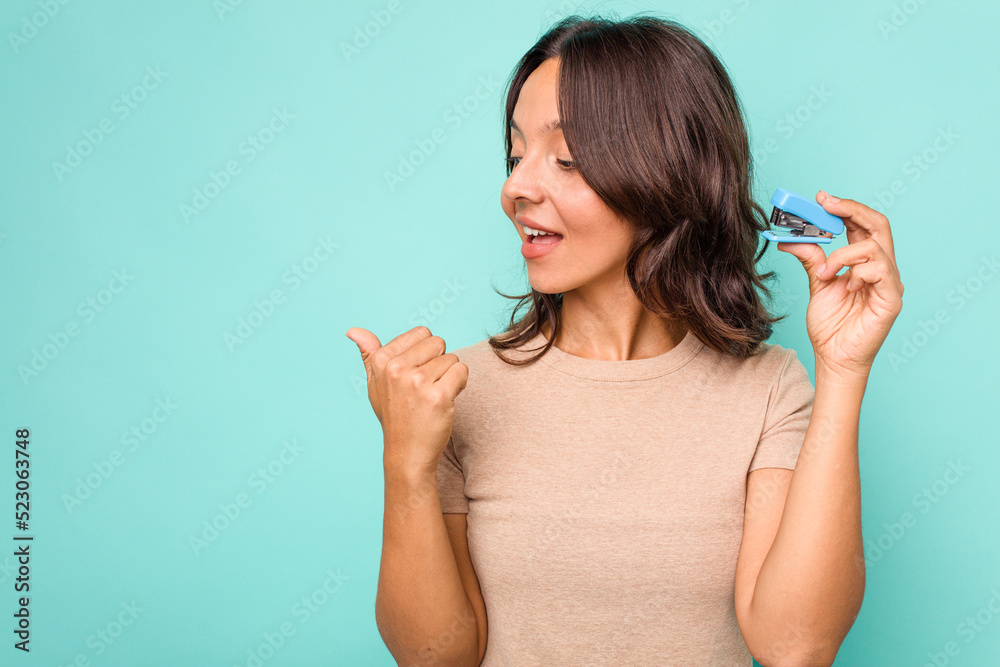 Young hispanic woman holding stapler isolated on blue background points with thumb finger away, laughing and carefree. Young hispanic woman holding stapler isolated on blue background points with thumb finger away, laughing and carefree.