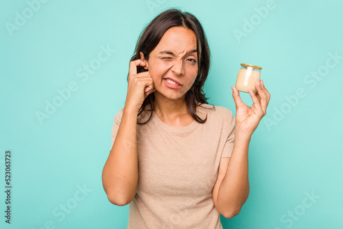 Cuadro en lienzo Young hispanic woman holding yogurt isolated on blue background covering ears with hands