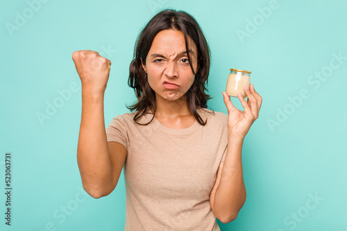Fotografia Young hispanic woman holding yogurt isolated on blue background showing fist to camera, aggressive facial expression