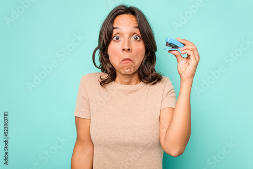 Young hispanic woman holding stapler isolated on blue background shrugs shoulders and open eyes confused.