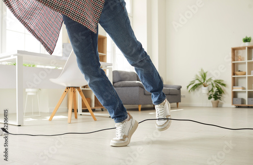 Man falls down as he stumbles over a power cable at home. Clumsy guy in sneakers trips over an electrical cord while walking on the floor in the living room. Cropped shot. Domestic accident concept