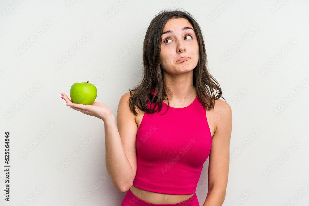 Young caucasian woman holding an apple isolated on white background confused, feels doubtful and unsure.
