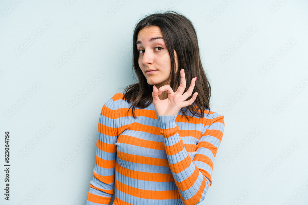 Young caucasian woman isolated on blue background winks an eye and holds an okay gesture with hand. Young caucasian woman isolated on blue background winks an eye and holds an okay gesture with hand.