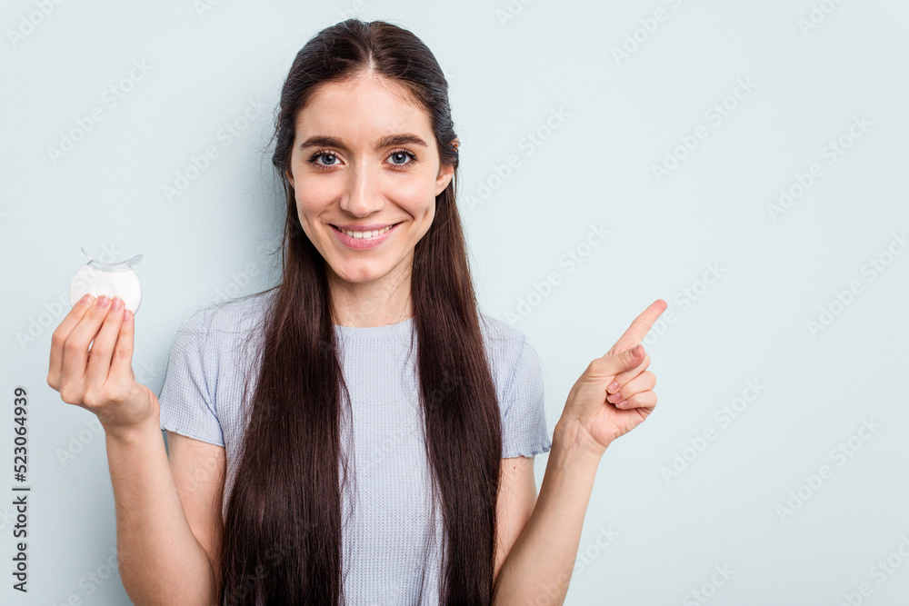 Young caucasian woman holding teeth whitener isolated on blue background smiling and pointing aside, showing something at blank space. Young caucasian woman holding teeth whitener isolated on blue background smiling and pointing aside, showing something at blank space.