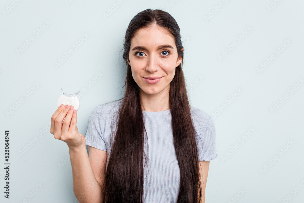 Young caucasian woman holding teeth whitener isolated on blue background happy, smiling and cheerful. Young caucasian woman holding teeth whitener isolated on blue background happy, smiling and cheerful.
