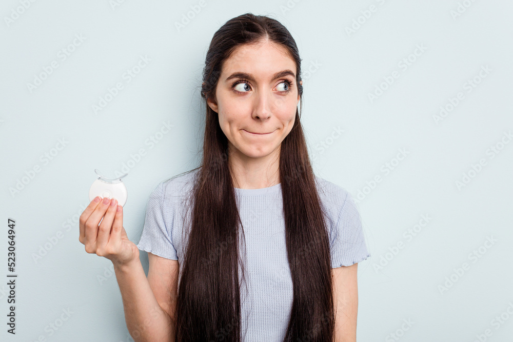 Young caucasian woman holding teeth whitener isolated on blue background confused, feels doubtful and unsure. Young caucasian woman holding teeth whitener isolated on blue background confused, feels doubtful and unsure.