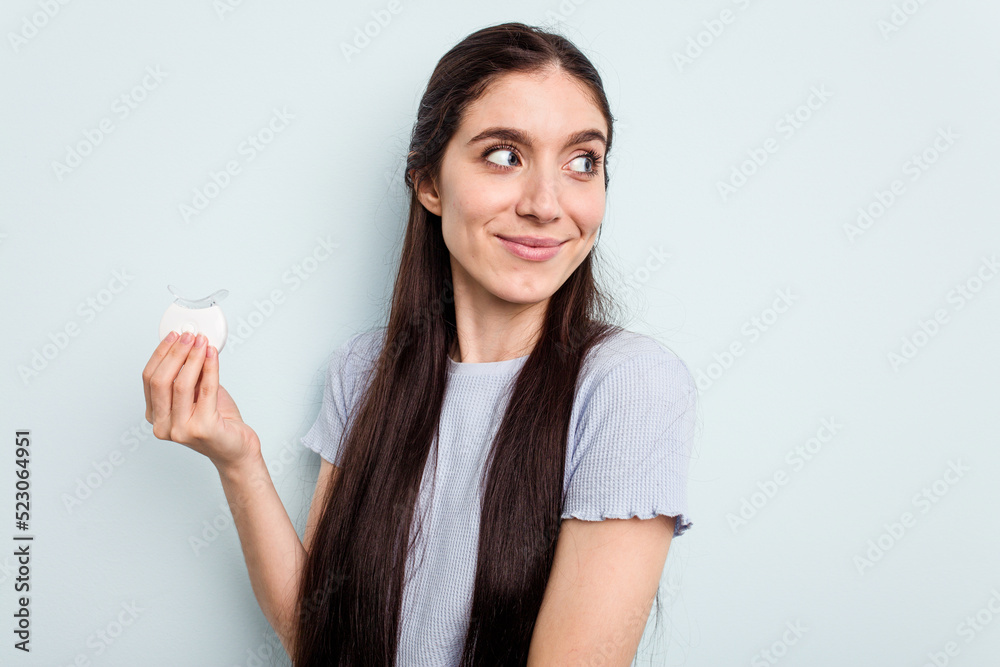 Young caucasian woman holding teeth whitener isolated on blue background looks aside smiling, cheerful and pleasant. Young caucasian woman holding teeth whitener isolated on blue background looks aside smiling, cheerful and pleasant.