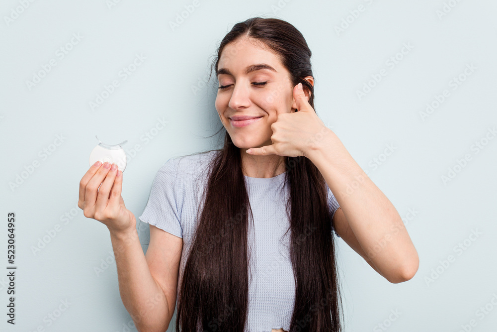 Young caucasian woman holding teeth whitener isolated on blue background showing a mobile phone call gesture with fingers. Young caucasian woman holding teeth whitener isolated on blue background showing a mobile phone call gesture with fingers.