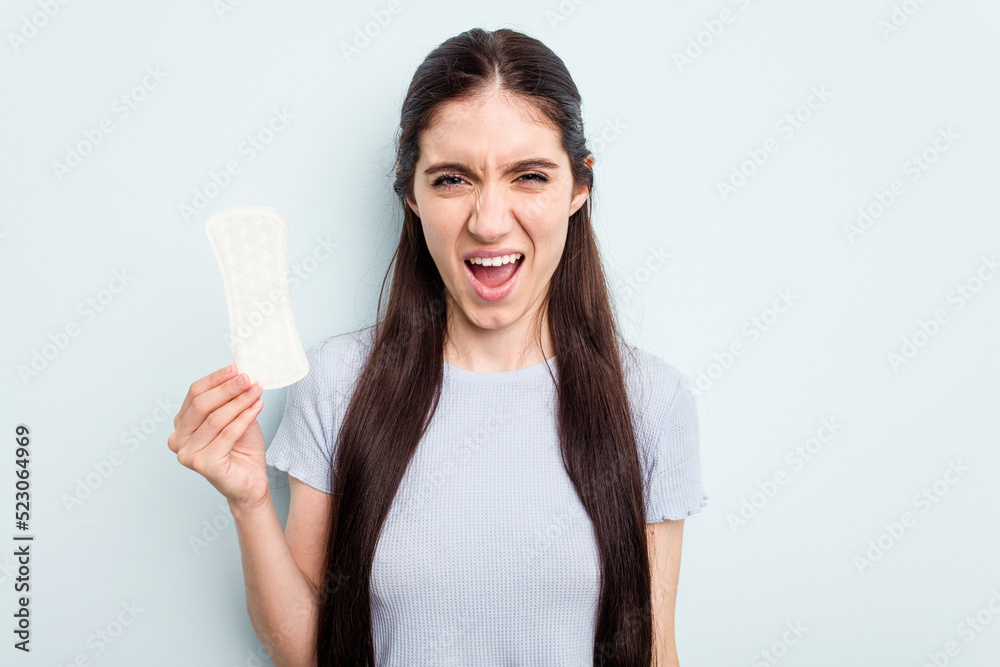 Young caucasian woman holding a sanitary napkin isolated on blue background screaming very angry and aggressive. Young caucasian woman holding a sanitary napkin isolated on blue background screaming very angry and aggressive.