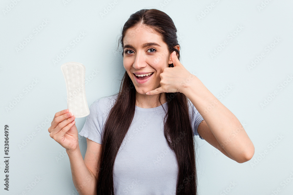 Young caucasian woman holding a sanitary napkin isolated on blue background showing a mobile phone call gesture with fingers. Young caucasian woman holding a sanitary napkin isolated on blue background showing a mobile phone call gesture with fingers.