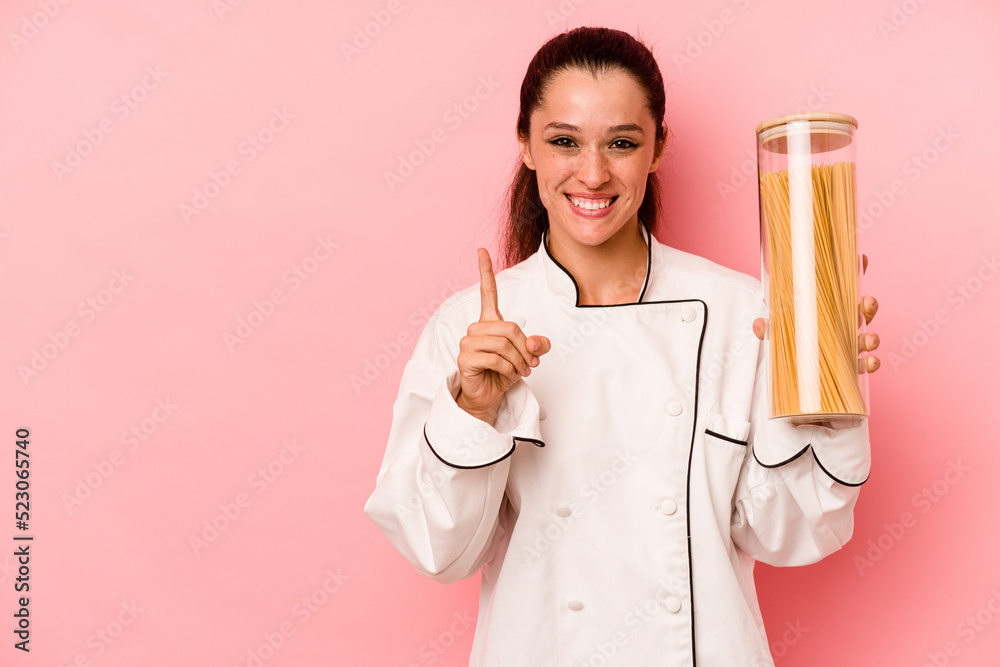 Young chef caucasian woman holding a spaghettis jar isolated on pink background showing number one with finger. Young chef caucasian woman holding a spaghettis jar isolated on pink background showing number one with finger.