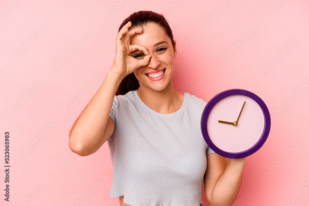 Young caucasian woman holding a clock isolated on pink background excited keeping ok gesture on eye. Young caucasian woman holding a clock isolated on pink background excited keeping ok gesture on eye.