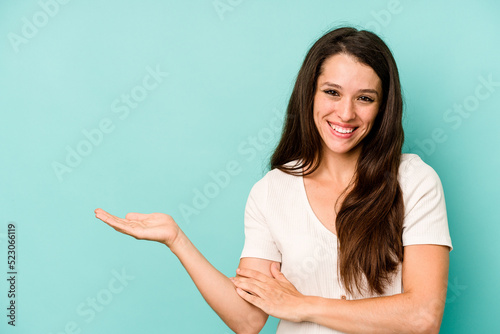 Young caucasian woman isolated on blue background showing a copy space on a palm and holding another hand on waist.