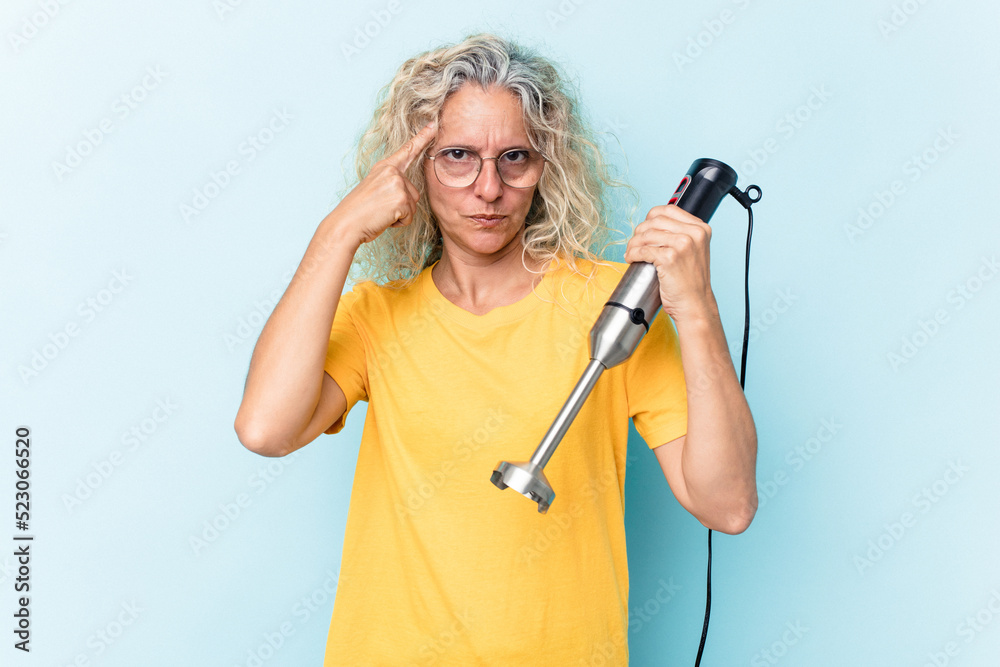 Middle age woman holding a blender isolated on blue background pointing temple with finger, thinking, focused on a task.