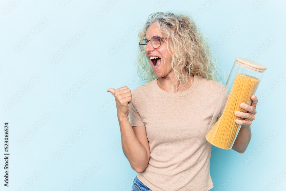 Middle age caucasian woman holding a spaghettis jar isolated on blue background points with thumb finger away, laughing and carefree. Middle age caucasian woman holding a spaghettis jar isolated on blue background points with thumb finger away, laughing and carefree.