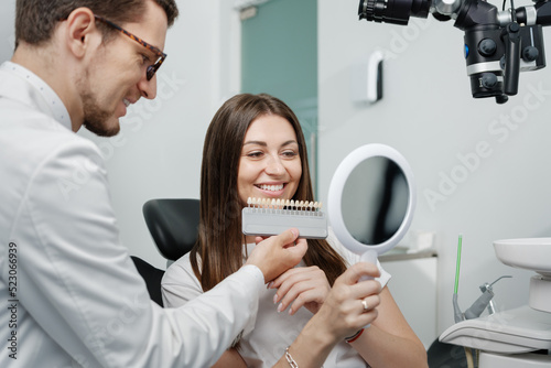 Young man dentist in white coat show veneers enamel color palette for beautiful patient woman