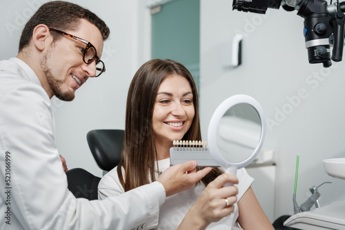Young man dentist in white coat show veneers enamel color palette for beautiful patient woman photo