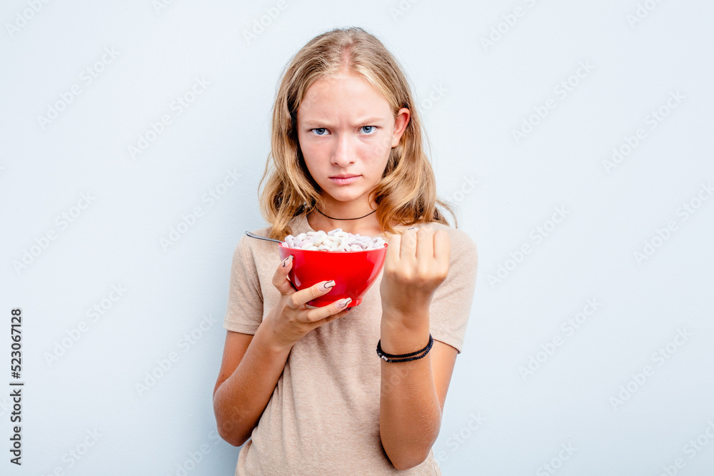 Caucasian teen girl eating cereals isolated on blue background showing fist to camera, aggressive facial expression. Caucasian teen girl eating cereals isolated on blue background showing fist to camera, aggressive facial expression.