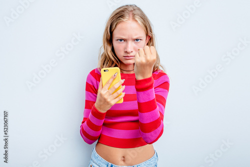 Caucasian teen girl holding mobile phone isolated on blue background showing fist to camera, aggressive facial expression.