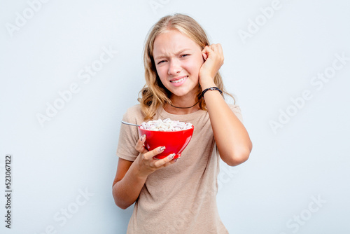 Caucasian teen girl eating cereals isolated on blue background covering ears with hands. © Asier