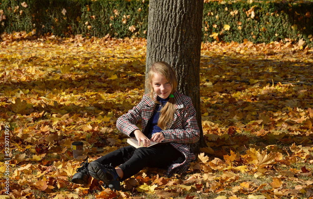 a beautiful girl sits leaning against a tree trunk on fallen leaves with a book and a cup of coffee or tea in an autumn park. a woman enjoys the warm rays of the autumn sun and relaxes a beautiful girl sits leaning against a tree trunk on fallen leaves with a book and a cup of coffee or tea in an autumn park. a woman enjoys the warm rays of the autumn sun and relaxes