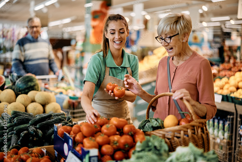 Happy sales clerk assists senior woman in buying vegetable at supermarket.