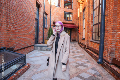 blonde with lilac strands of hair in a beige coat and glasses on the background of a red brick building