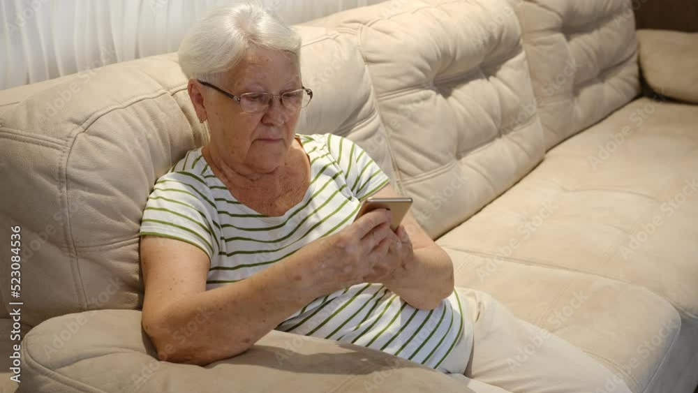 Pleasant senior older lady resting on couch, using applications on smartphone. Happy old mature woman chatting in messenger or social network with friends or children, shopping in internet store Pleasant senior older lady resting on couch, using applications on smartphone. Happy old mature woman chatting in messenger or social network with friends or children, shopping in internet store
