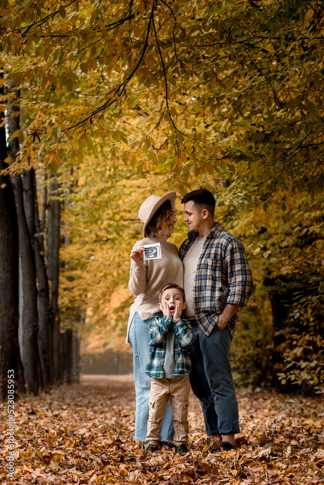 Happy family of a man with a woman holding an ultrasound pregnancy picture and a surprised son who will be the older brother. Autumn photo shoot of a family waiting to be replenished