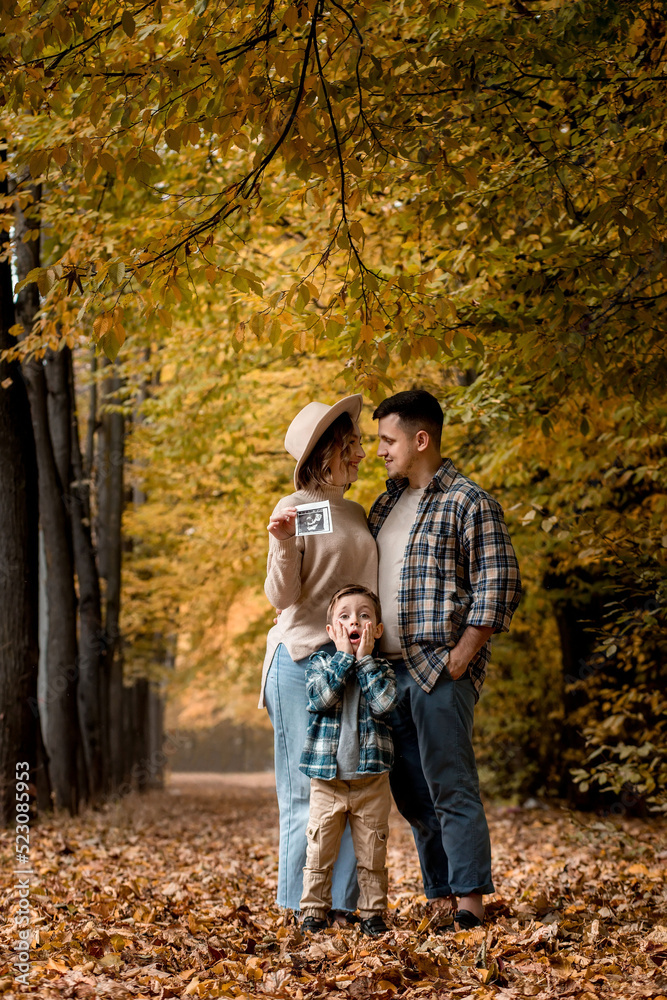 Happy family of a man with a woman holding an ultrasound pregnancy picture and a surprised son who will be the older brother. Autumn photo shoot of a family waiting to be replenished Happy family of a man with a woman holding an ultrasound pregnancy picture and a surprised son who will be the older brother. Autumn photo shoot of a family waiting to be replenished