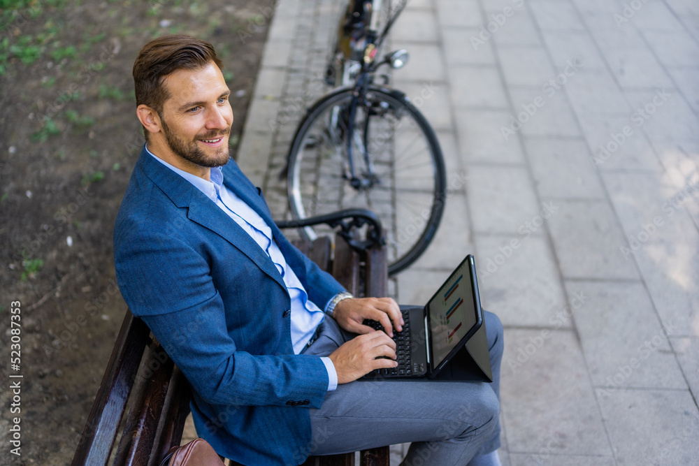 Obraz premium Portrait of happy entrepreneur working on laptop\tablet while sitting on park bench. Bicycle in blurred background.
