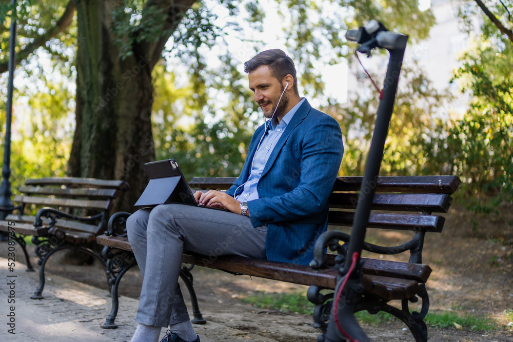 Caucasian male entrepreneur freelancer sitting outdoors in city park a bench talking online a video call using laptop or tablet. Business man office worker working on urban street background