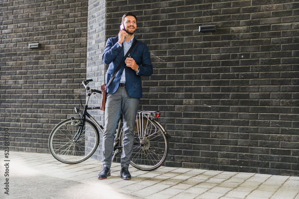 Caucasian men in suit standing in front of his bicycle, he using his phone and drinking morning coffee Caucasian men in suit standing in front of his bicycle, he using his phone and drinking morning coffee
