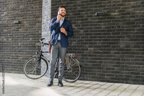 Caucasian men in suit standing in front of his bicycle, he using his phone and drinking morning coffee