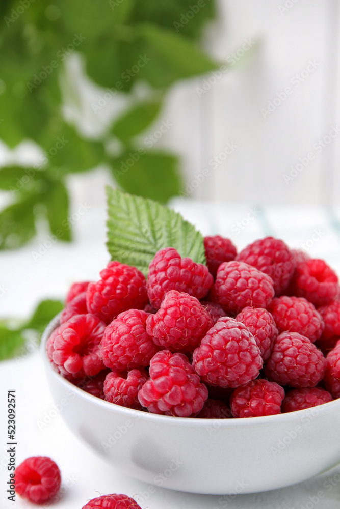 Bowl of fresh ripe raspberries with green leaf on white table against blurred background, closeup Bowl of fresh ripe raspberries with green leaf on white table against blurred background, closeup