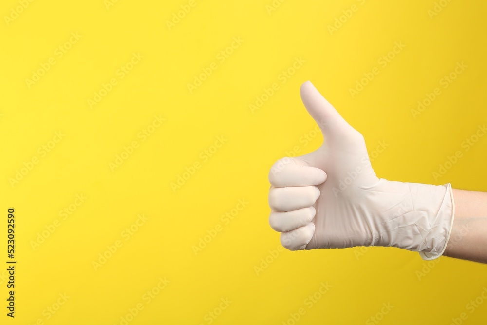Person in medical gloves showing thumb up on yellow background, closeup of hand. Space for text