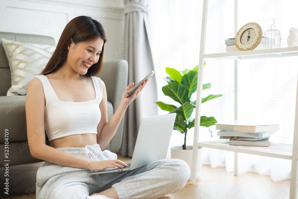 Freelance asian woman in glasses with mobile phone typing at laptop and working from home office. Happy girl sitting on couch in living room. Freelance asian woman in glasses with mobile phone typing at laptop and working from home office. Happy girl sitting on couch in living room.