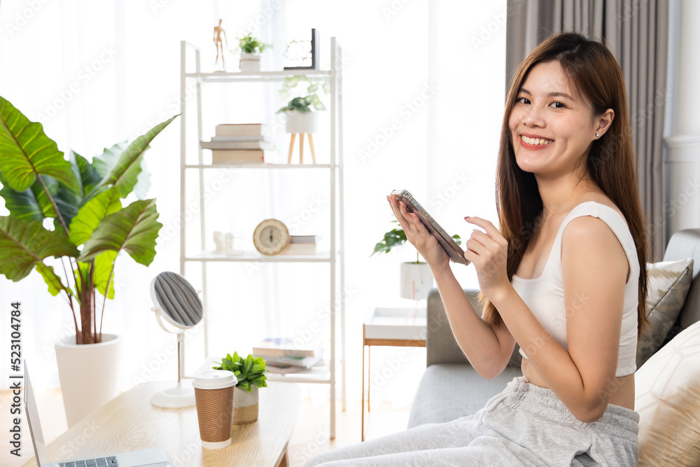 Happy and cheerful woman is using a phone sitting on a sofa.Asian young woman looking mobile phone celebrating with good news or discount voucher for shopping online at home. Happy and cheerful woman is using a phone sitting on a sofa.Asian young woman looking mobile phone celebrating with good news or discount voucher for shopping online at home.