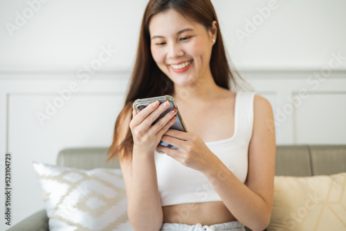 Happy and cheerful woman is using a phone sitting on a sofa.Asian young woman looking mobile phone celebrating with good news or discount voucher for shopping online at home.