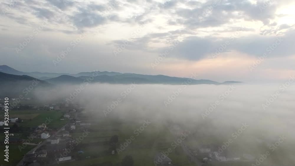 Paisaje aéreo de dron sobrevolando pueblo rural en el campo cubierto por niebla al atardecer, con las montañas de fondo sobre las nubes Paisaje aéreo de dron sobrevolando pueblo rural en el campo cubierto por niebla al atardecer, con las montañas de fondo sobre las nubes