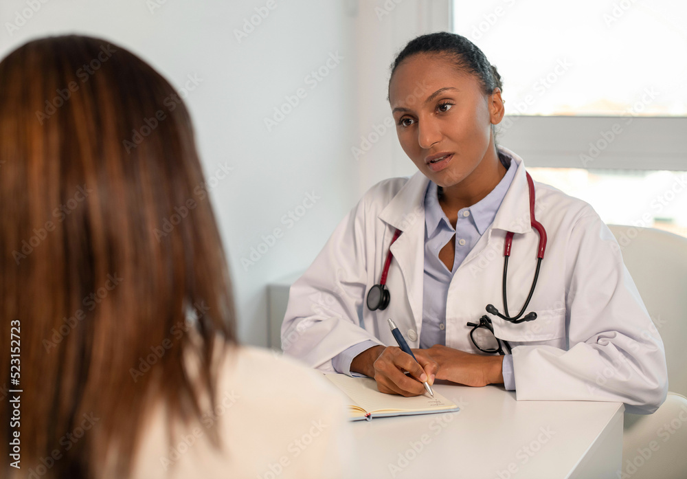 Fototapeta premium Confident young doctor writing down anamnesis of female patient. African American general practitioner sitting at table and talking to woman in her office. Visit to doctor concept