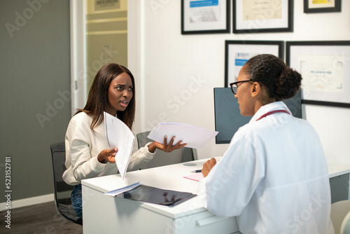 Angry young female patient arguing with doctor about contract. African American woman sitting at doctors office exasperated with documents. Medical services contract, medical insurance concept