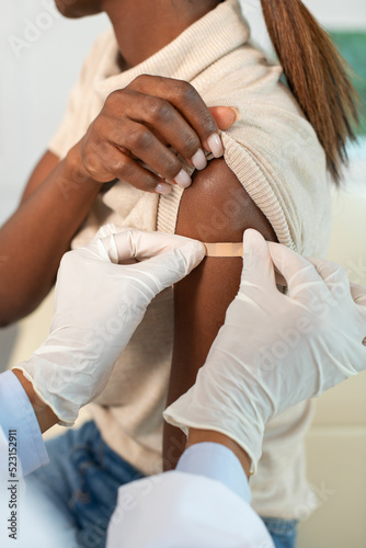 Close-up of hands of nurse in protective gloves applying plaster to female arm. Doctor putting patch on shoulder of patient after injection. Vaccination and immunization concept