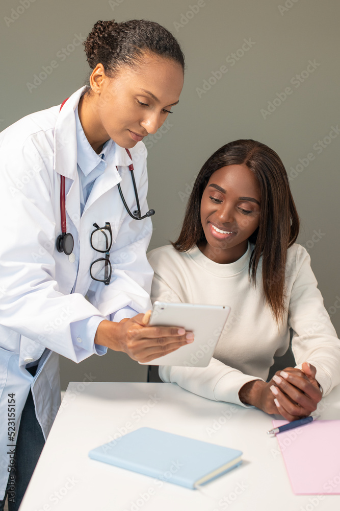Fototapeta premium Happy woman looking at positive medical tests on digital tablet. Confident young African American doctor consulting female patient in clinic. Medical consultation concept