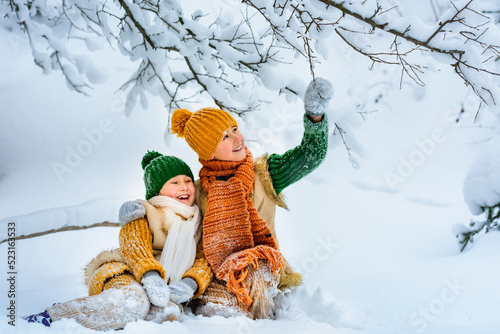 Two cute little cheerful children are playing with snow among snowdrifts. Children sit in the snow under snow-covered tree. Winter activities for children. Knitted scarf, hat, sweater, woolen coat.