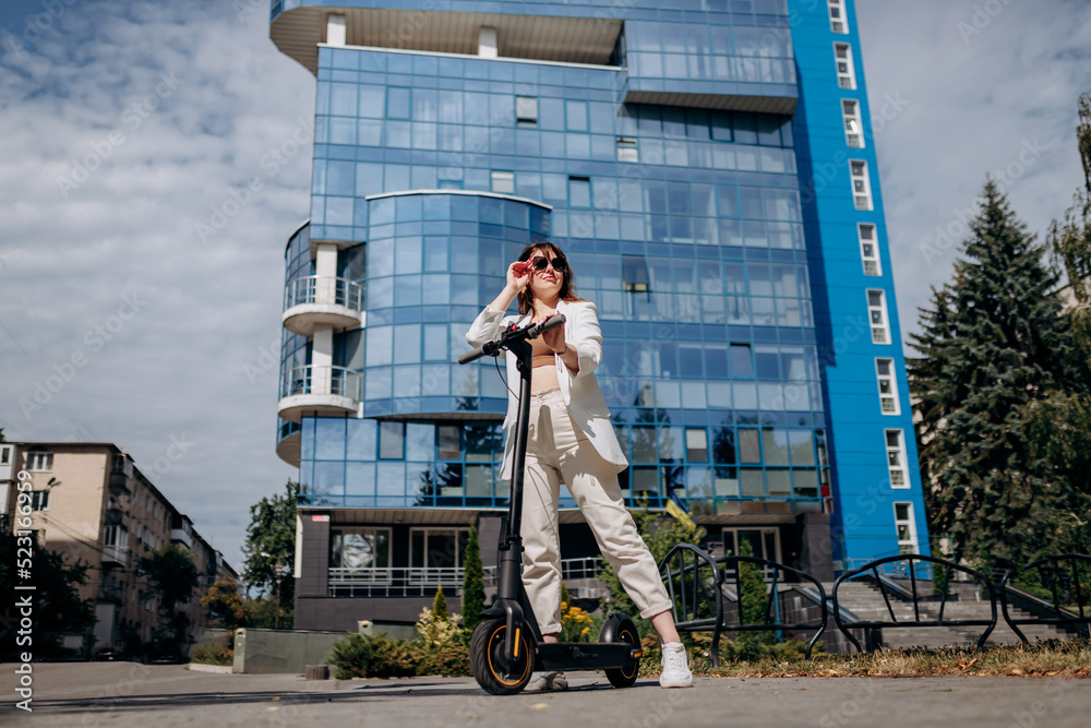 Beautiful young woman in sunglasses and white suit standing on her electric scooter near modern building and looking away