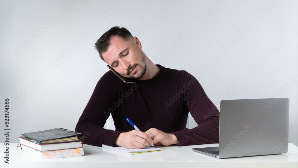 A man talks on the phone and records information in the office at his desk.